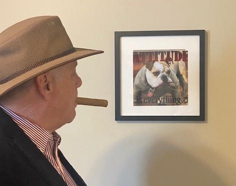 Mark Melrose in a hat with a cigar, looking at a framed print reading Attitude is everything.